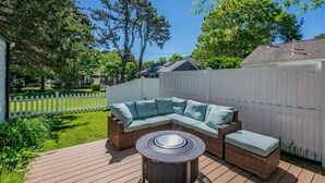 A deck with a wicker sectional sofa and a round table, surrounded by a white picket fence and lush greenery.