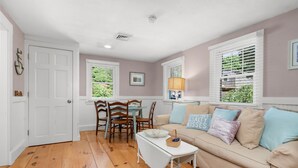 A cozy living room with a beige sofa, wooden dining table, and chairs. There are two windows with blinds, a white door, and a ceiling light.