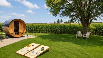 Eine holzartige, fassförmige Struktur mit einer Tür, zwei Adirondack-Stühlen und einem Maisfeld im Hintergrund.