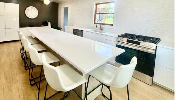 A modern kitchen with a long white countertop, white chairs, a stove, and a clock on the wall.
