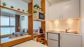 Eine kompakte Küchenzeile mit Mikrowelle, Spüle und Kühlschrank. Ein kleiner Esstisch und ein Stuhl sind vorhanden. Das Zimmer verfügt über Holzregale mit Pflanzen und ein Fenster mit Vorhängen.