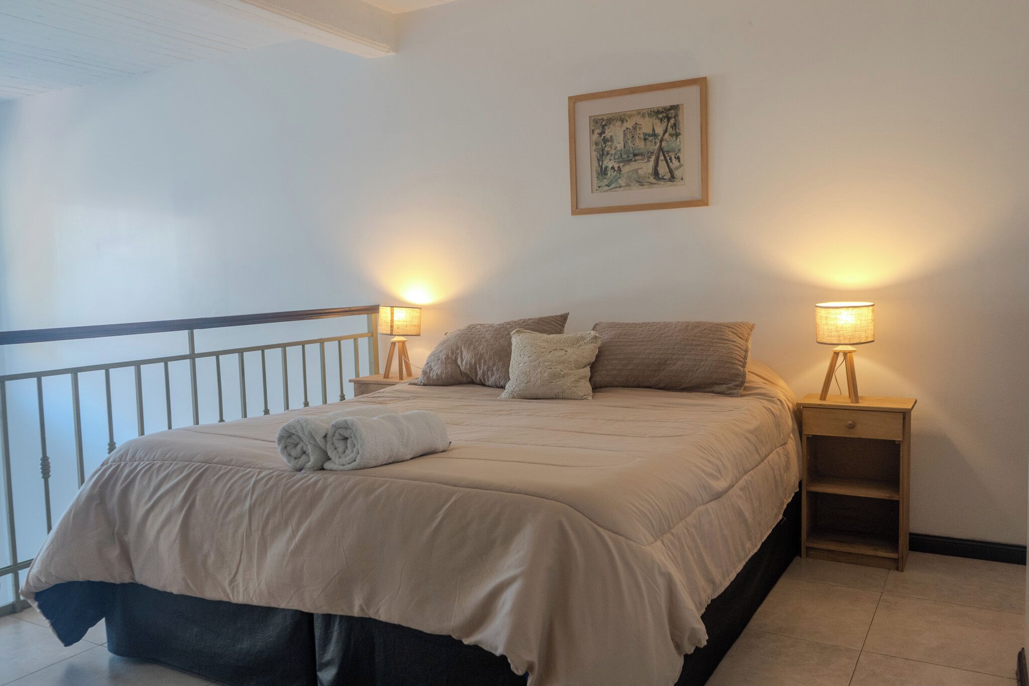 Una cama bien hecha con una colcha color beige, dos lámparas de noche, un cuadro en la pared y una baranda de balcón.