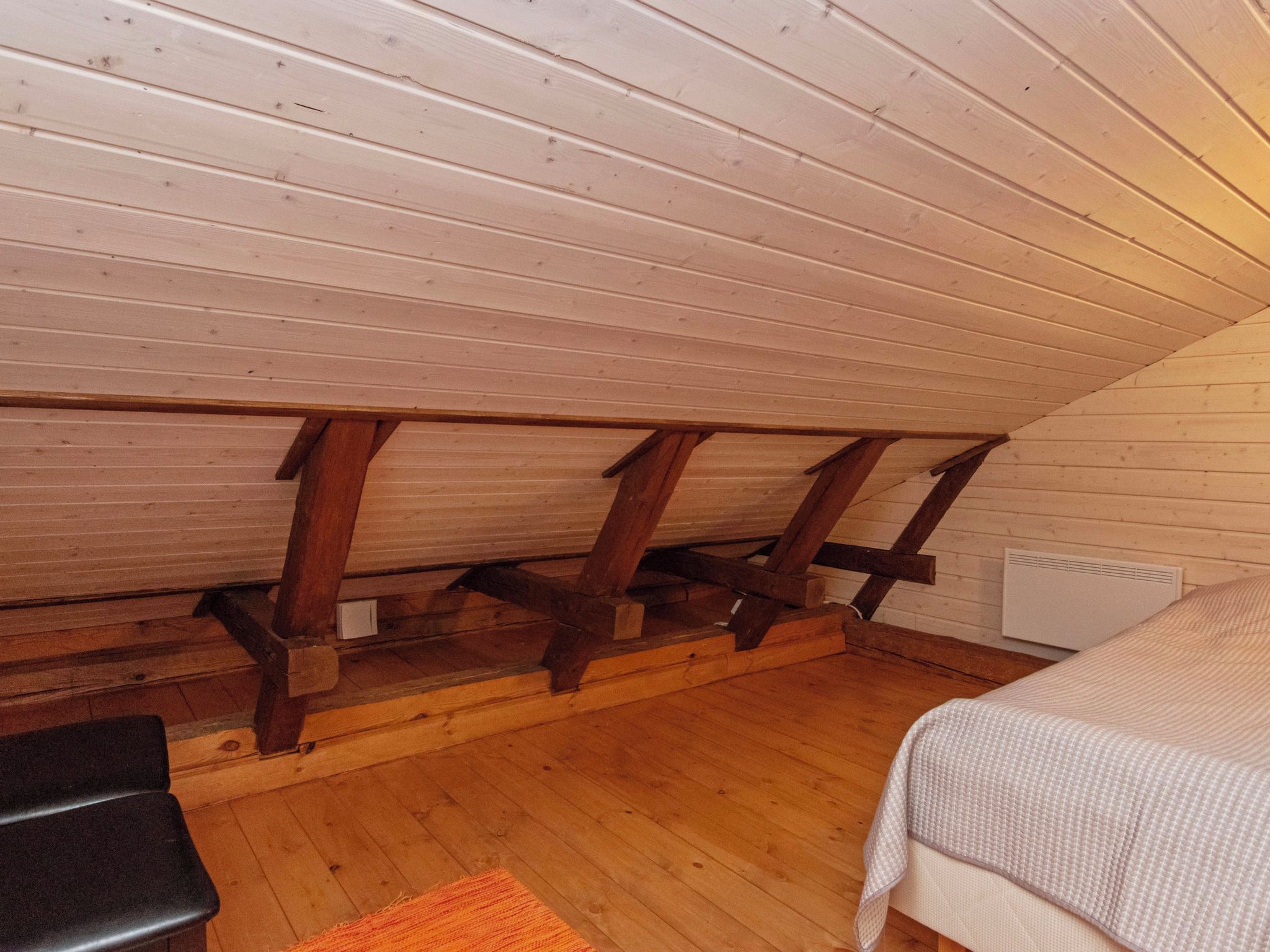 Quarto com teto inclinado, vigas de madeira, cama, mesa pequena e radiador.