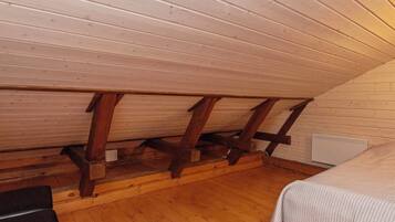 Quarto com teto inclinado, vigas de madeira, cama, mesa pequena e radiador.