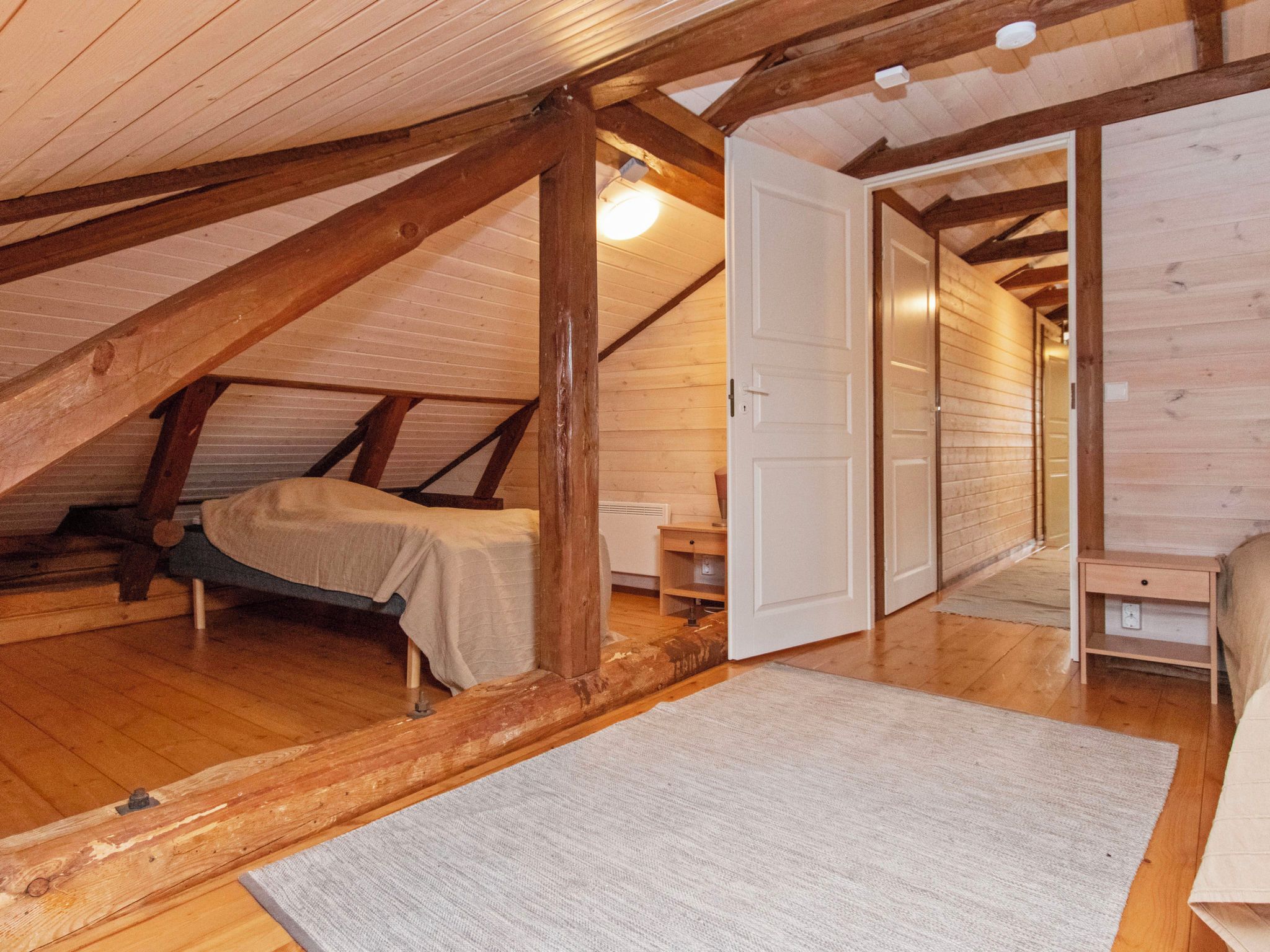 A loft bedroom with a sloped ceiling, wooden beams, a bed, a nightstand, and a door leading to another room.