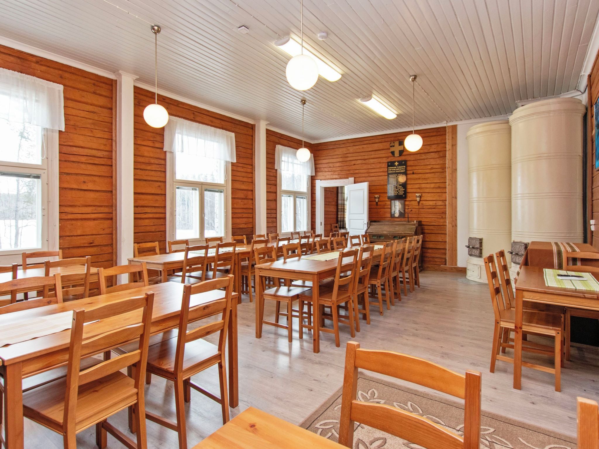 Uma sala de jantar com mesas e cadeiras de madeira, janelas amplas e teto branco.