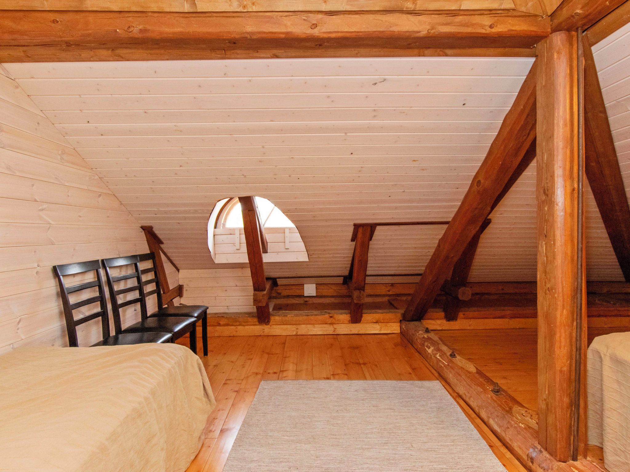 Ein Zimmer mit schräger Decke, Holzbalken, einem Fenster, einem Bett, einem Sessel und einem Teppich.