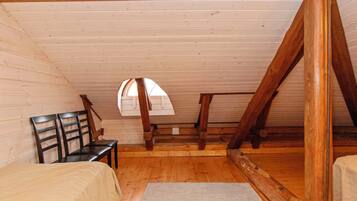 Ein Zimmer mit schräger Decke, Holzbalken, einem Fenster, einem Bett, einem Sessel und einem Teppich.