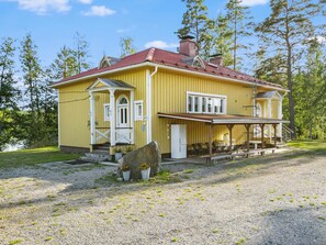 A yellow house with a red roof, a porch, and a stone in front.