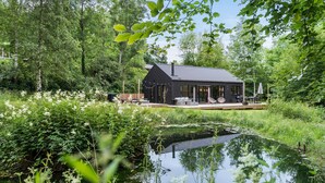 Una moderna casa nera con veranda, immersa nel verde e con uno stagno.
