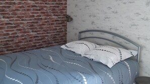 Een enkel bed met een metalen frame, een blauw beddegoed met een patroon, en een wit kussen met stipjes. De kamer heeft aan de ene kant een bakstenen muur en aan de andere kant behang met een patroon.