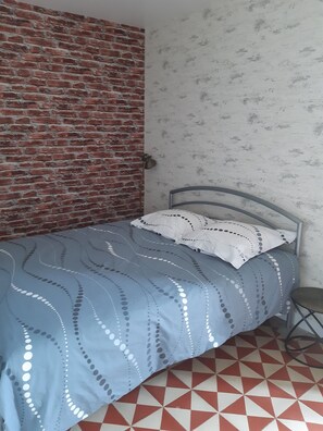 Een enkel bed met een metalen frame, een blauw beddegoed met een patroon, en een wit kussen met stipjes. De kamer heeft aan de ene kant een bakstenen muur en aan de andere kant behang met een patroon.