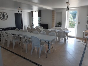 Een ruime eetruimte met witte tafels en stoelen, een grote klok aan de muur, en een deur naar buiten.