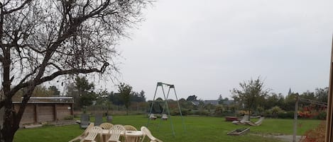 Uma área de pátio com mesa e cadeiras, balanço e jardim com gramado e árvores.