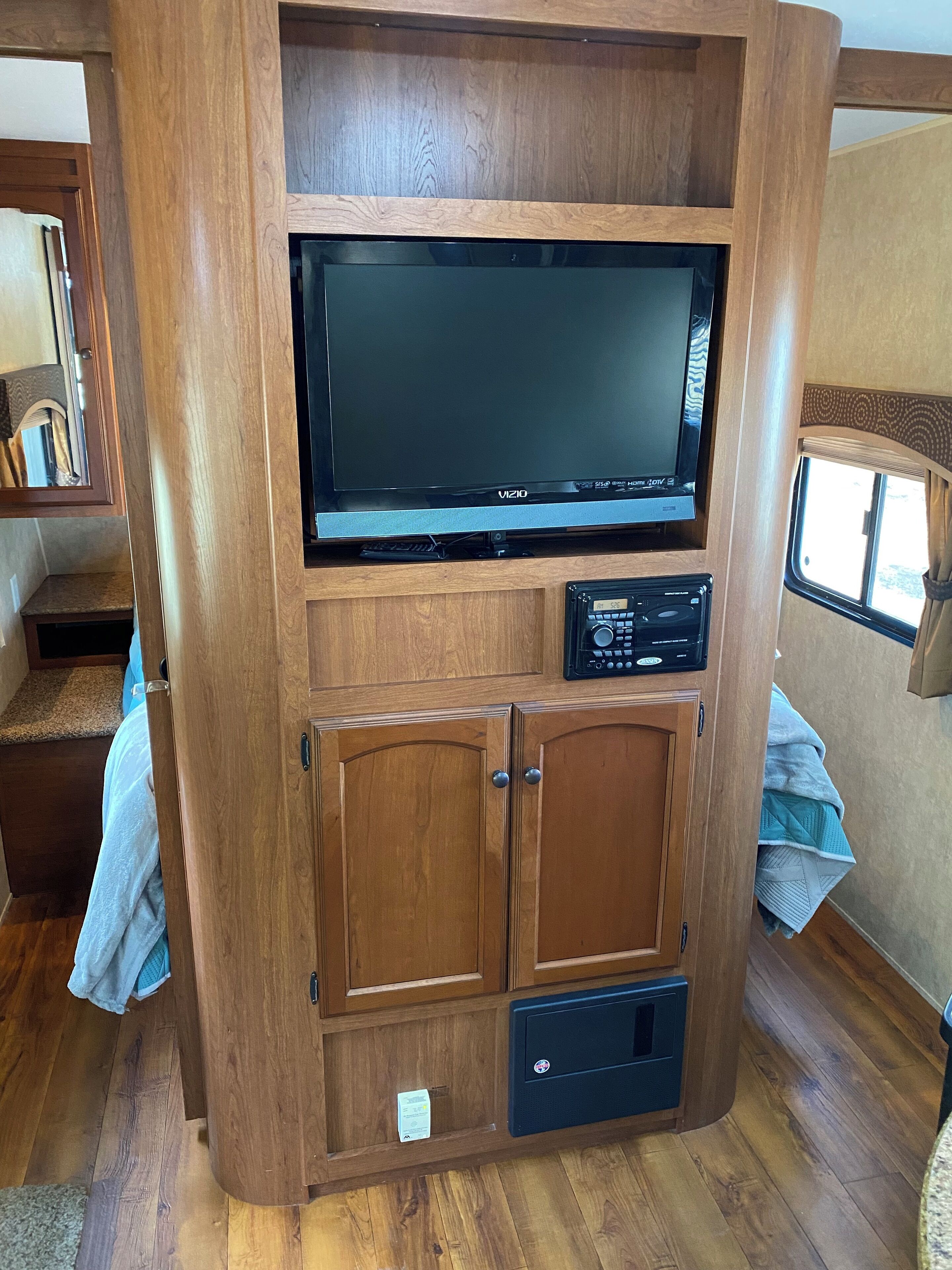 A compact RV interior with a large flat-screen TV mounted on a wooden cabinet, a built-in radio, and a small storage compartment.