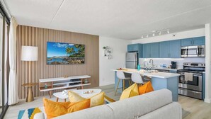 A modern living room with a sofa, a dining area, and a kitchen with blue cabinets and stainless steel appliances.
