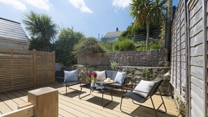 A wooden deck with two black lounge chairs and a small table, surrounded by stone walls and greenery.