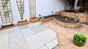 Un jardín con jardineras elevadas, una estructura para plantas trepadoras y una zona de estar con sillas.