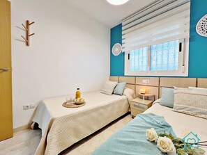 A bedroom with two beds, a bedside table with a lamp, a window with blinds, and a wall-mounted coat rack.