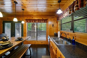 A cabin interior with a wooden dining table, bench, kitchen area, and a view through the door. - Catch A Good Time · Scenic Escape: Pet Friendly, Hot Tub & Fire Pit (Broken Bow)