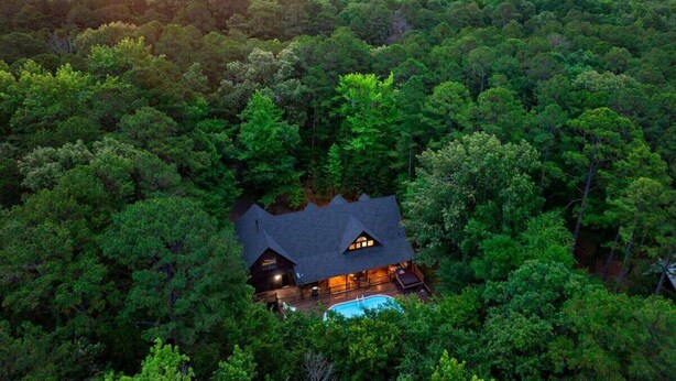 A secluded cabin surrounded by dense forest, with a swimming pool and a well-lit interior. - Wild Oasis · New! Wild Oasis: Pool, Gameroom, Theatre, Hot Tub (Broken Bow)