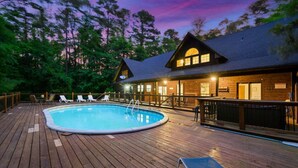 A wooden deck with a pool, lounge chairs, and a building with a large window and a porch. - Wild Oasis · New! Wild Oasis: Pool, Gameroom, Theatre, Hot Tub (Broken Bow)