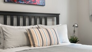 Una cama bien hecha con almohadas a rayas y lisas, un cabecero oscuro y una planta pequeña al lado de una lámpara sobre una pared blanca.