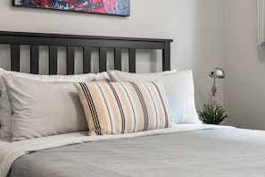 Un lit bien fait, avec des oreillers à rayures et unis, une tête de lit sombre et une petite plante placée à côté d’une lampe sur un mur blanc.