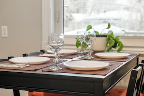 Une table à manger dressée avec des assiettes, des verres et des couverts, agrémentée d’une plante en pot et avec une vue sur la fenêtre en arrière-plan.