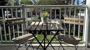 Una mesa de madera y dos sillas en un balcón con barandilla blanca, una planta en maceta y vistas a árboles y un garaje al fondo.