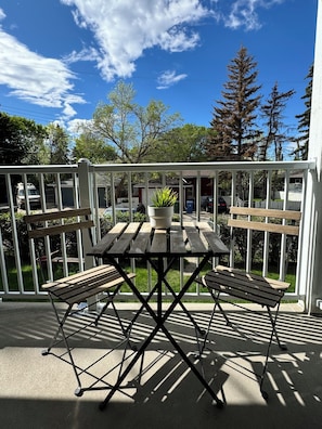 Une table en bois et deux chaises sur un balcon doté d’une balustrade blanche, un pot de fleurs et, en arrière-plan, une vue sur des arbres et un garage.