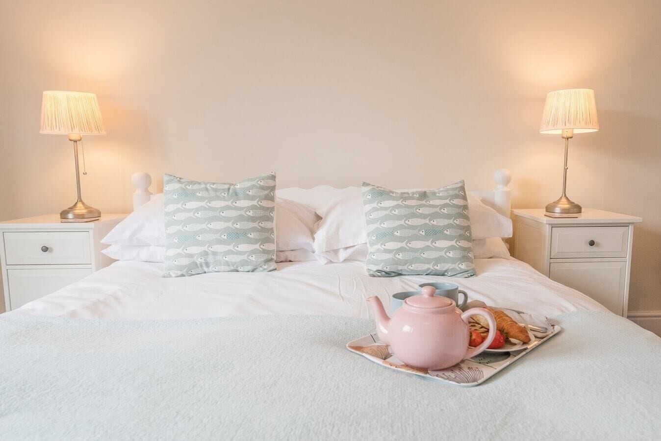 A neatly made bed with white bedding, two pillows with a fish pattern, and a pink teapot on a tray with a cup and a strawberry.