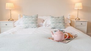 A neatly made bed with white bedding, two pillows with a fish pattern, and a pink teapot on a tray with a cup and a strawberry.