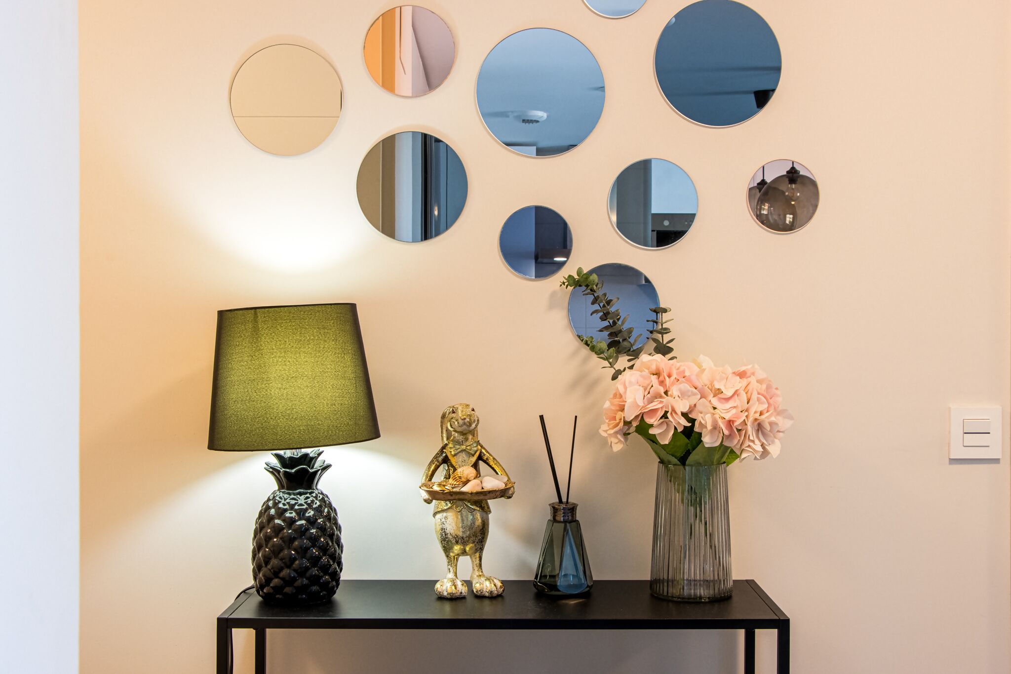Un mur orné de plusieurs miroirs ronds, ainsi qu’une petite table sur laquelle se trouvent une lampe, une statuette de grenouille décorative et un vase rempli de fleurs.