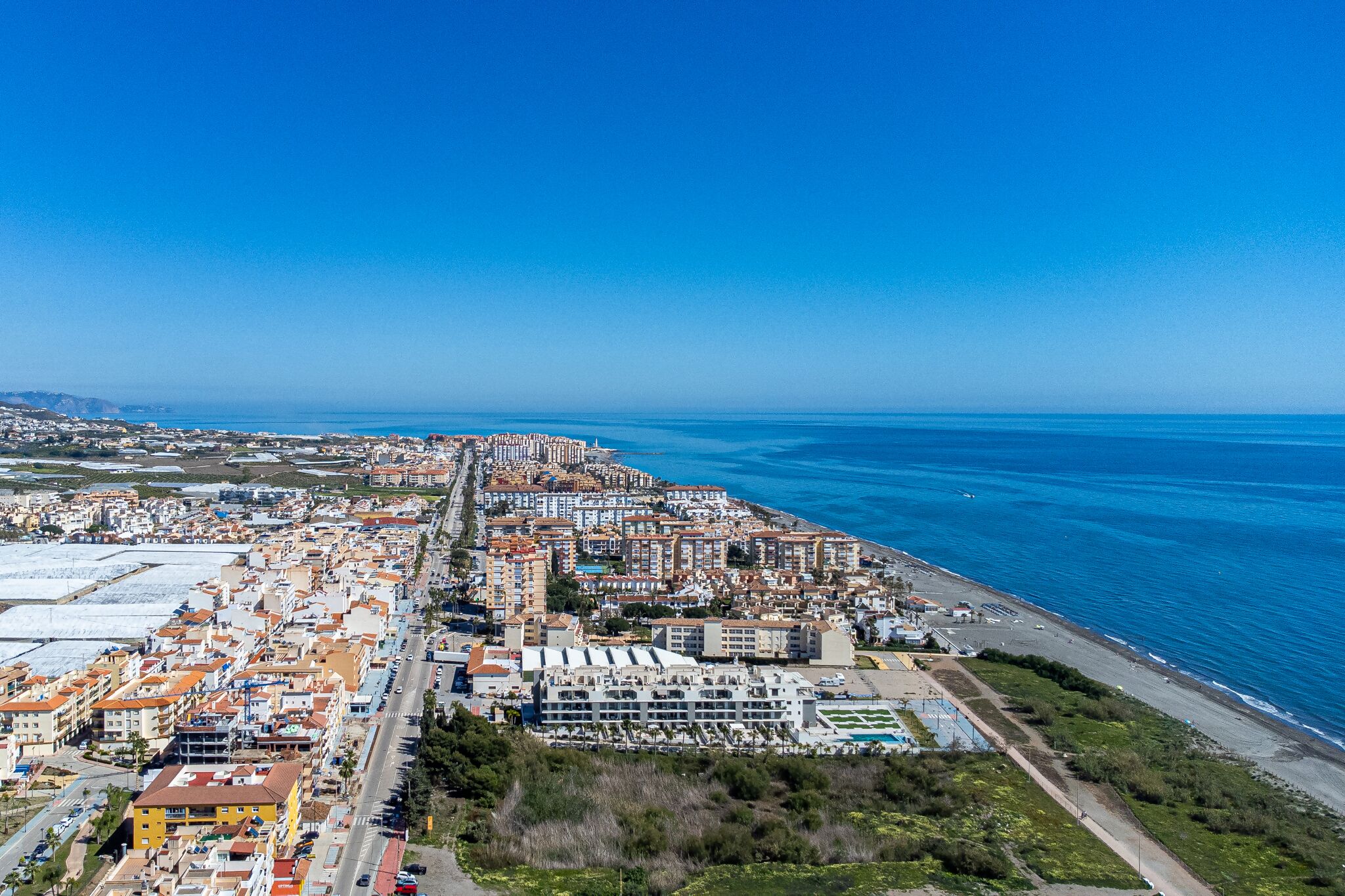 Vue aérienne d’une ville côtière, avec une longue plage, des immeubles d’habitation denses et un ciel bleu dégagé.