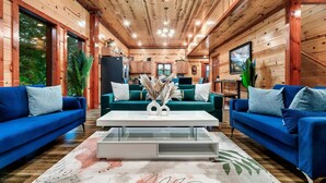 A log cabin interior with a living room featuring two blue sofas, a white coffee table, and a large window.