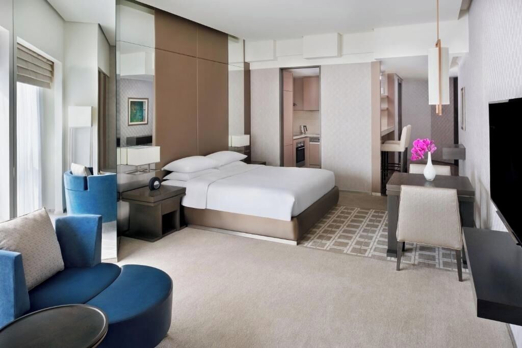 Une chambre d’hôtel moderne avec un grand lit, un coin salon doté d’un canapé bleu, une petite table, une télévision et un coin cuisine visible à travers une porte ouverte.