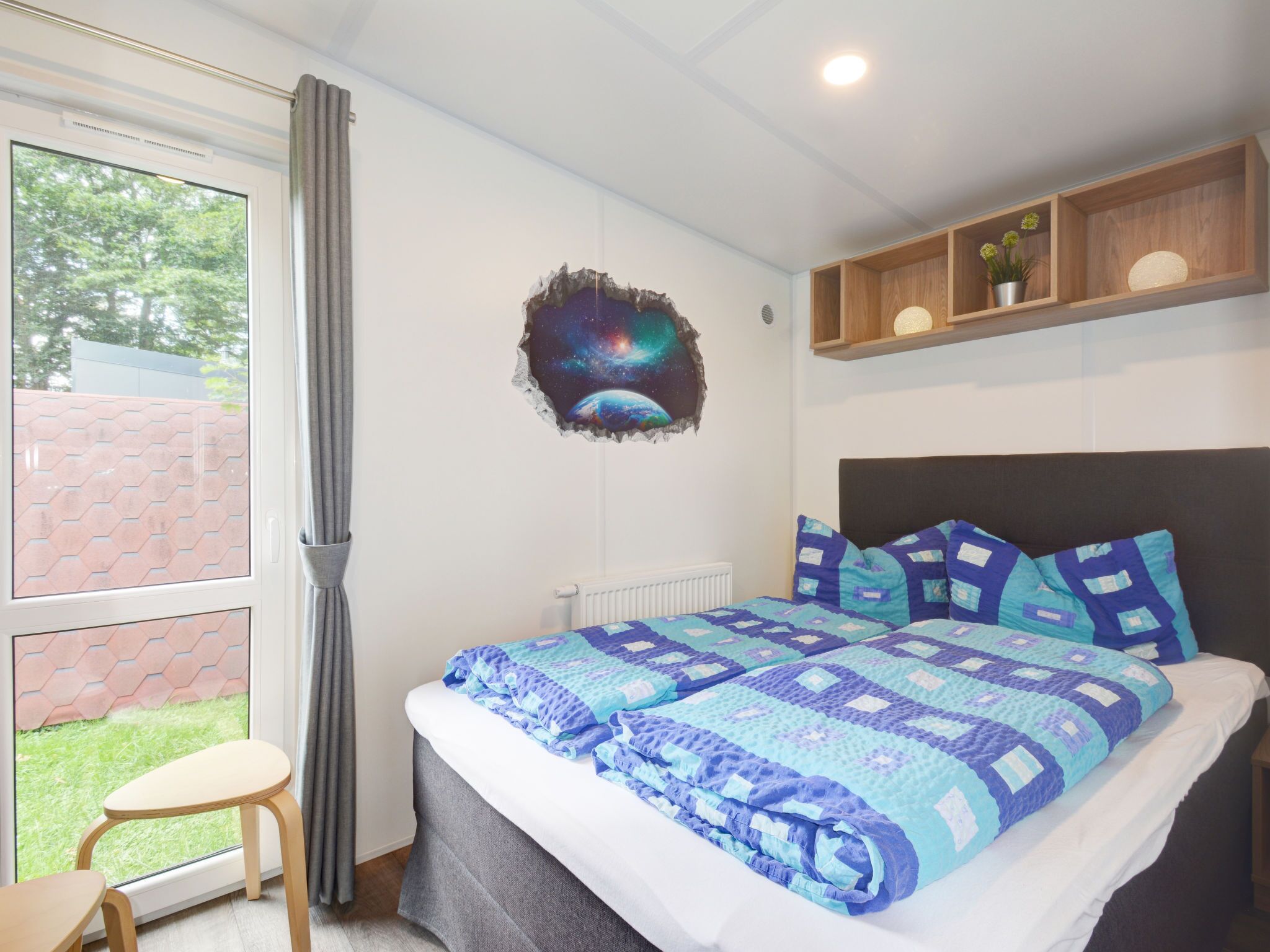 A bedroom with a large bed, a window with a view of a brick wall and greenery, a wall decoration, and a shelf with decorative items.