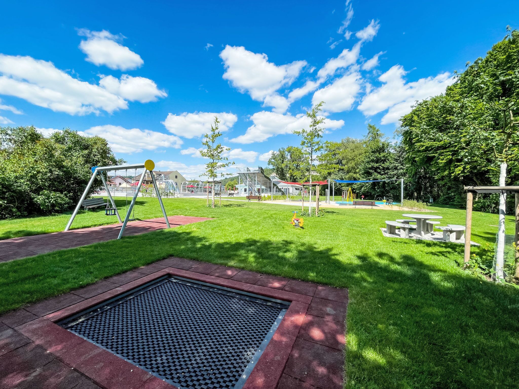 Une aire de jeux avec des balançoires et un petit toboggan, entourée d’herbe et d’arbres.