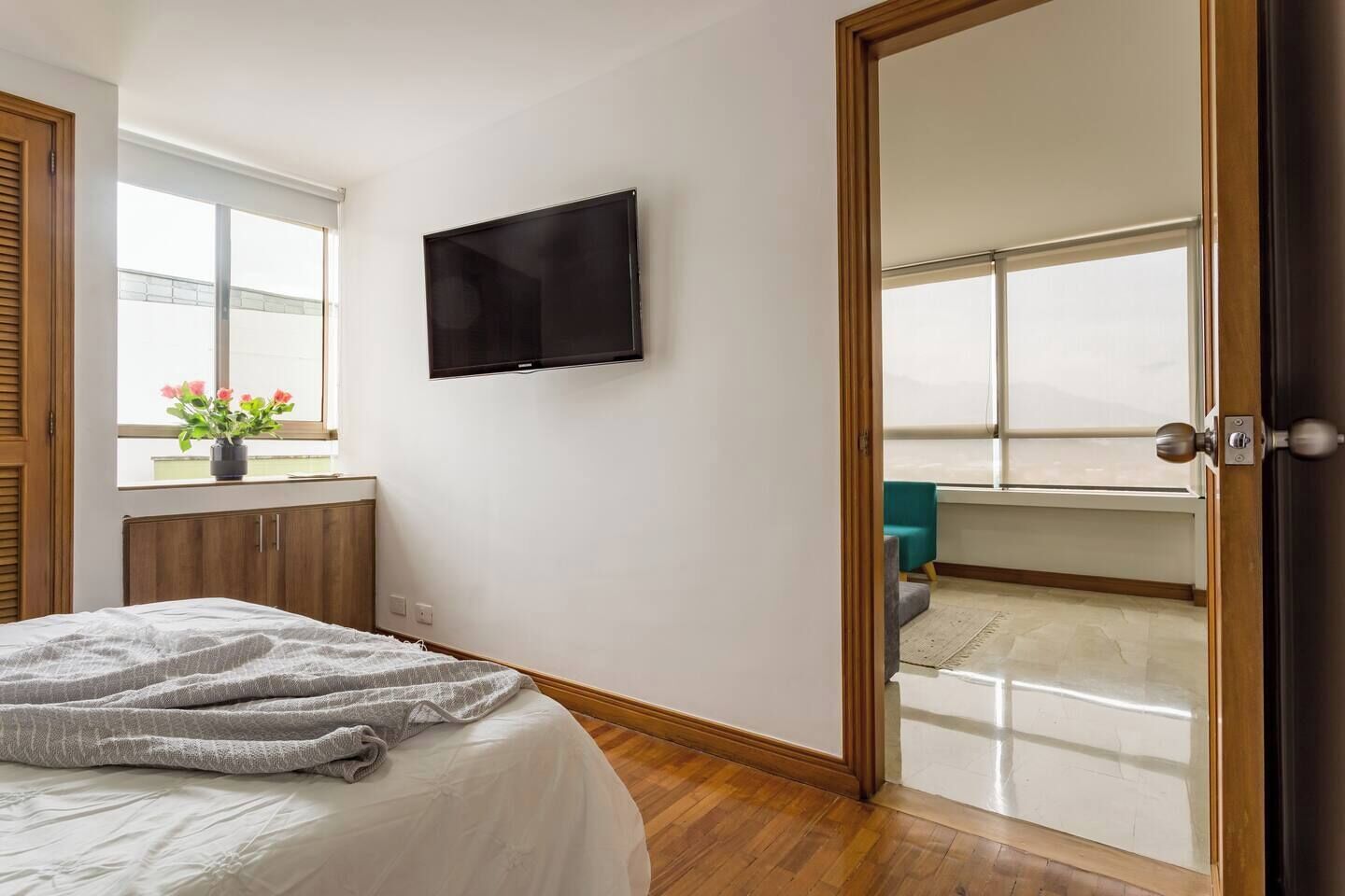Un dormitorio con cama, televisor montado en la pared, ventana con flores y puerta corredera a un balcón.