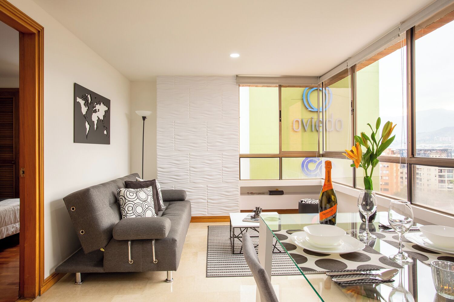 A modern living room with a grey sofa, a glass dining table, and a view of the city through large windows.