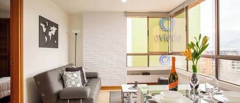 A modern living room with a grey sofa, a glass dining table, and a view of the city through large windows.