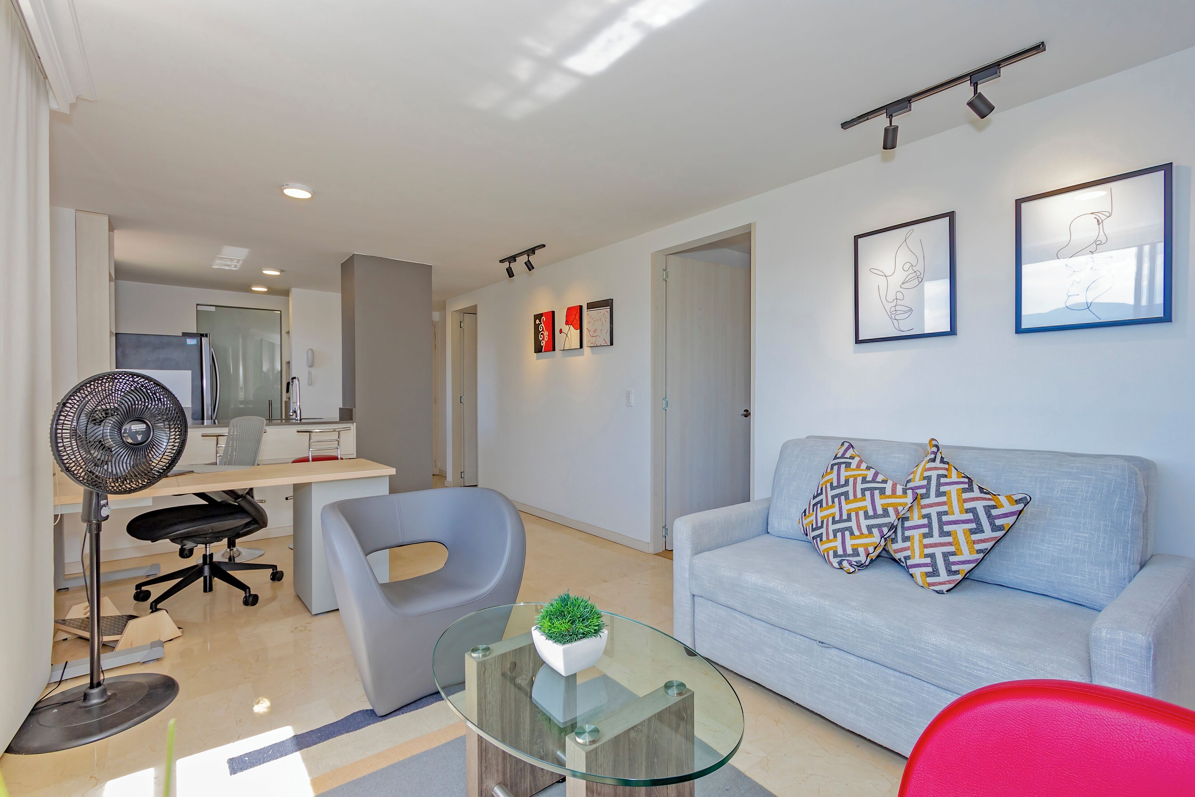 Uma sala de estar moderna com uma mesa de centro de vidro, um sofá cinza com almofadas estampadas, um ventilador de chão e uma escrivaninha com uma cadeira.