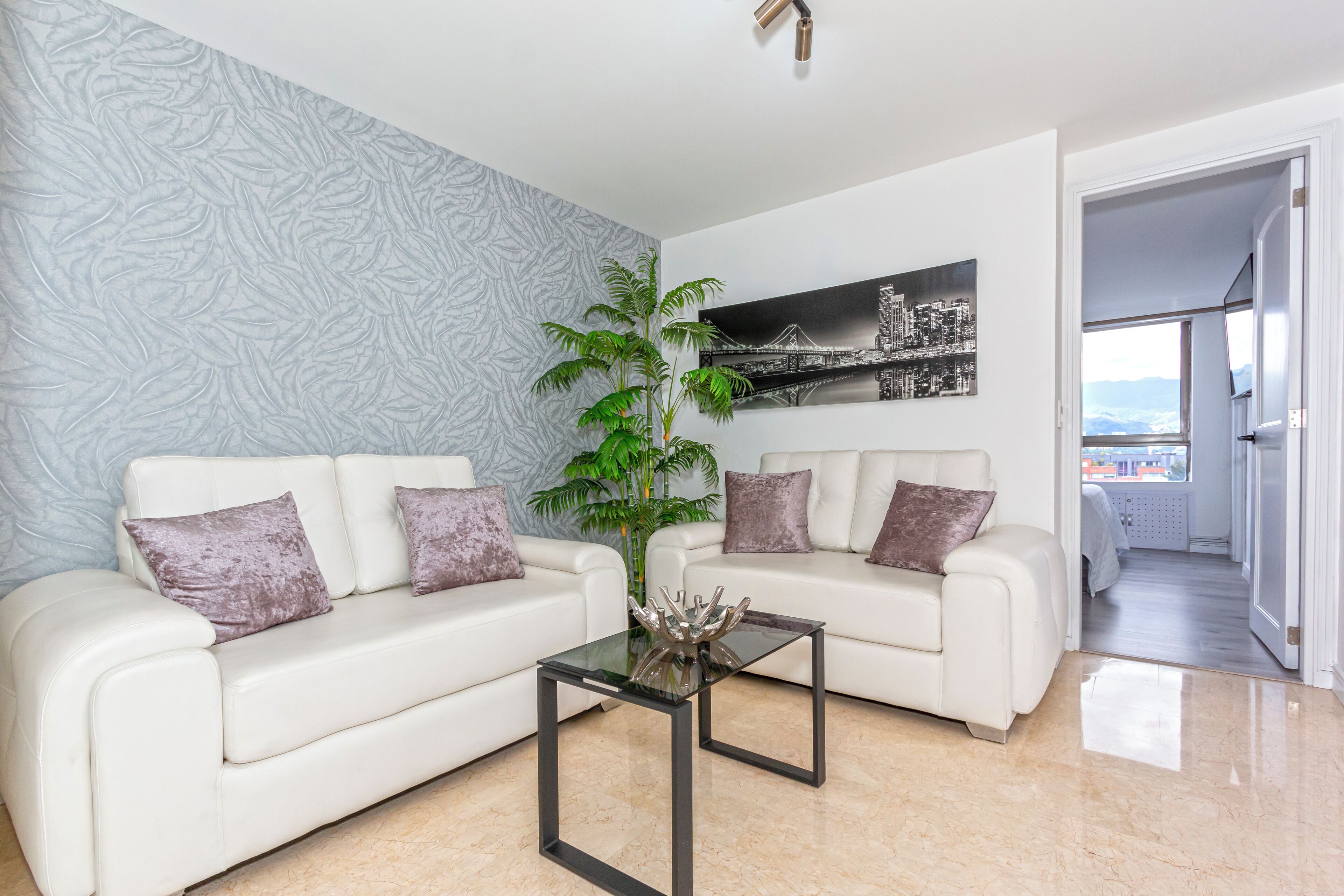 Un salon moderne avec deux canapés blancs, une table basse en verre et une grande plante en pot. Un tableau représentant un paysage urbain est accroché au mur, et une porte donne sur un balcon avec vue.
