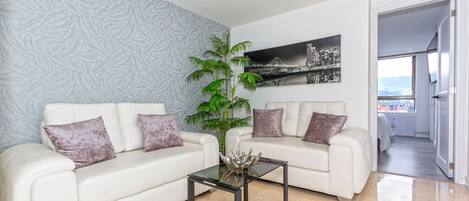 Un salon moderne avec deux canapés blancs, une table basse en verre et une grande plante en pot. Un tableau représentant un paysage urbain est accroché au mur, et une porte donne sur un balcon avec vue.