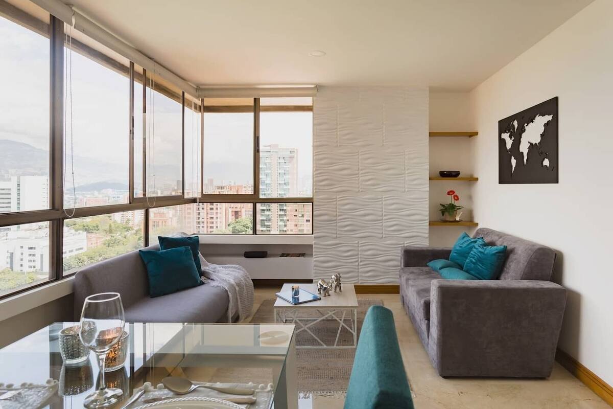 A modern living room with a glass dining table, two grey sofas with teal cushions, a world map artwork on the wall, and a view of city buildings through large windows.