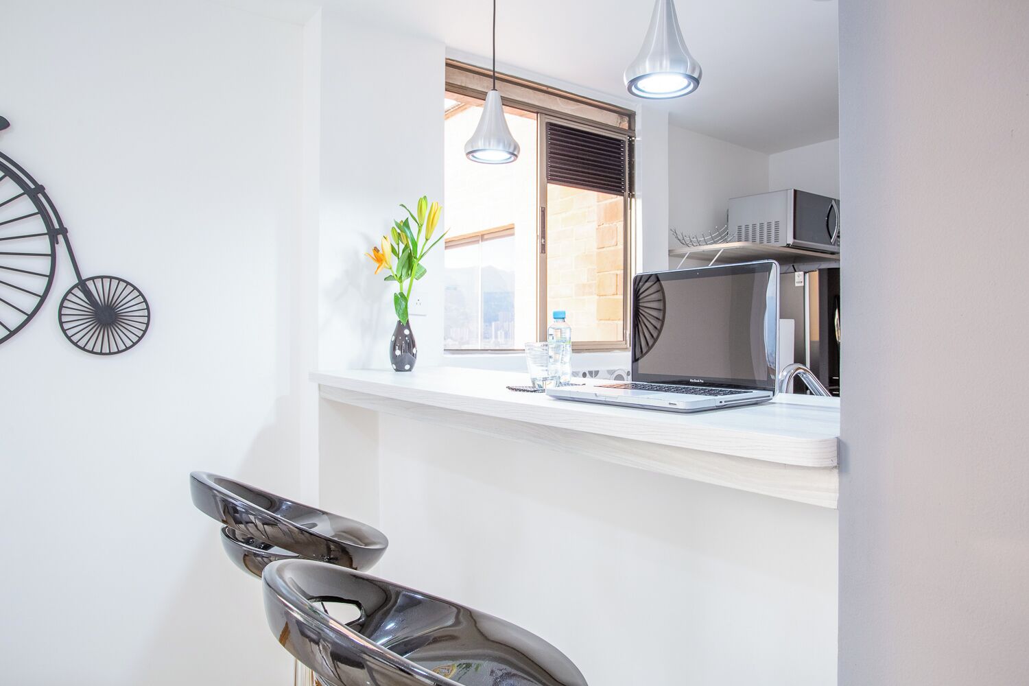 Una cocina moderna con encimera blanca, una laptop, un microondas y una ventana con persianas.