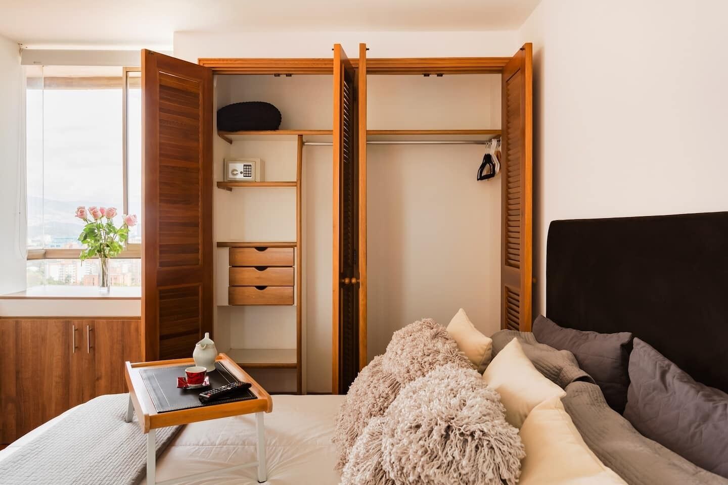 A bedroom with a bed, a bedside table with a cup and remote, a fluffy throw blanket, a wardrobe with a mirror, and a window with a view of buildings.