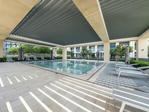 A covered outdoor swimming pool with lounge chairs and umbrellas, surrounded by greenery and modern buildings.