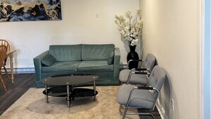 En stue med en grøn sofa, et sort sofabord og en grå lænestol. Der er et billede på væggen og et ur over sofaen.